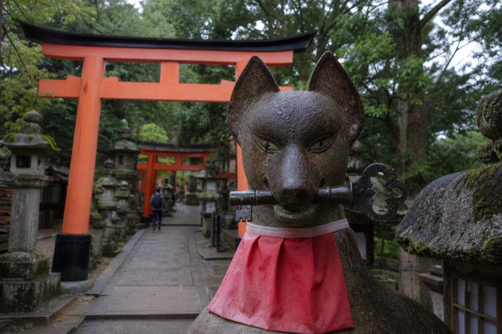 A stone fox statue holding a key in its mouth, framed by a red torii gate and moss-covered lanterns